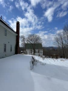 On the left is the back side of a white house with a tall brick chimney. In the center is a lot of hilly snow. There are leafless trees in the background.