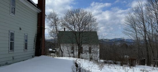 On the left is the back side of a white house with a tall brick chimney. In the center is a lot of hilly snow. There are leafless trees in the background.