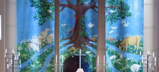 A church altar with a stunning large art piece behind it. The art is in three long banners, together they are forming a large tree with green leaves in front of a blue sky. There are animals and water flowing all around the roots.