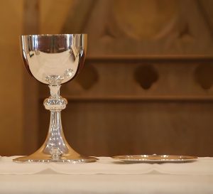 A silver chalice next to a small silver plate on a white tablecloth covered table with a wood wall in the background.