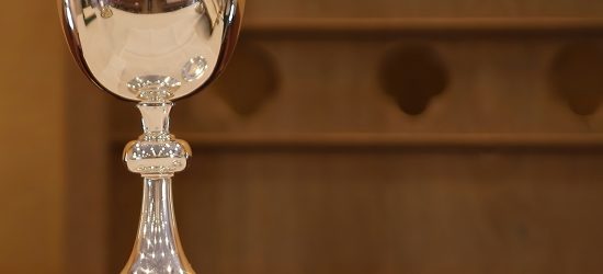 A silver chalice next to a small silver plate on a white tablecloth covered table with a wood wall in the background.