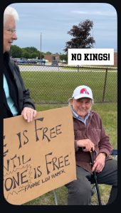 An elderly white man is sitting in a lawn chair, standing next to him is an elderly white woman holding a sign that is partially showing (it reads "No one is free until everyone is free"). They are at the No Kings protest.