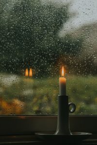 single candle lit and glowing by a window on a rainy lush green day