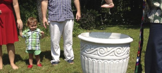 Outdoor baptism ceremony: an adult holds an infant over a large barrel used as a baptismal font, with onlookers gathered on a grassy lawn.